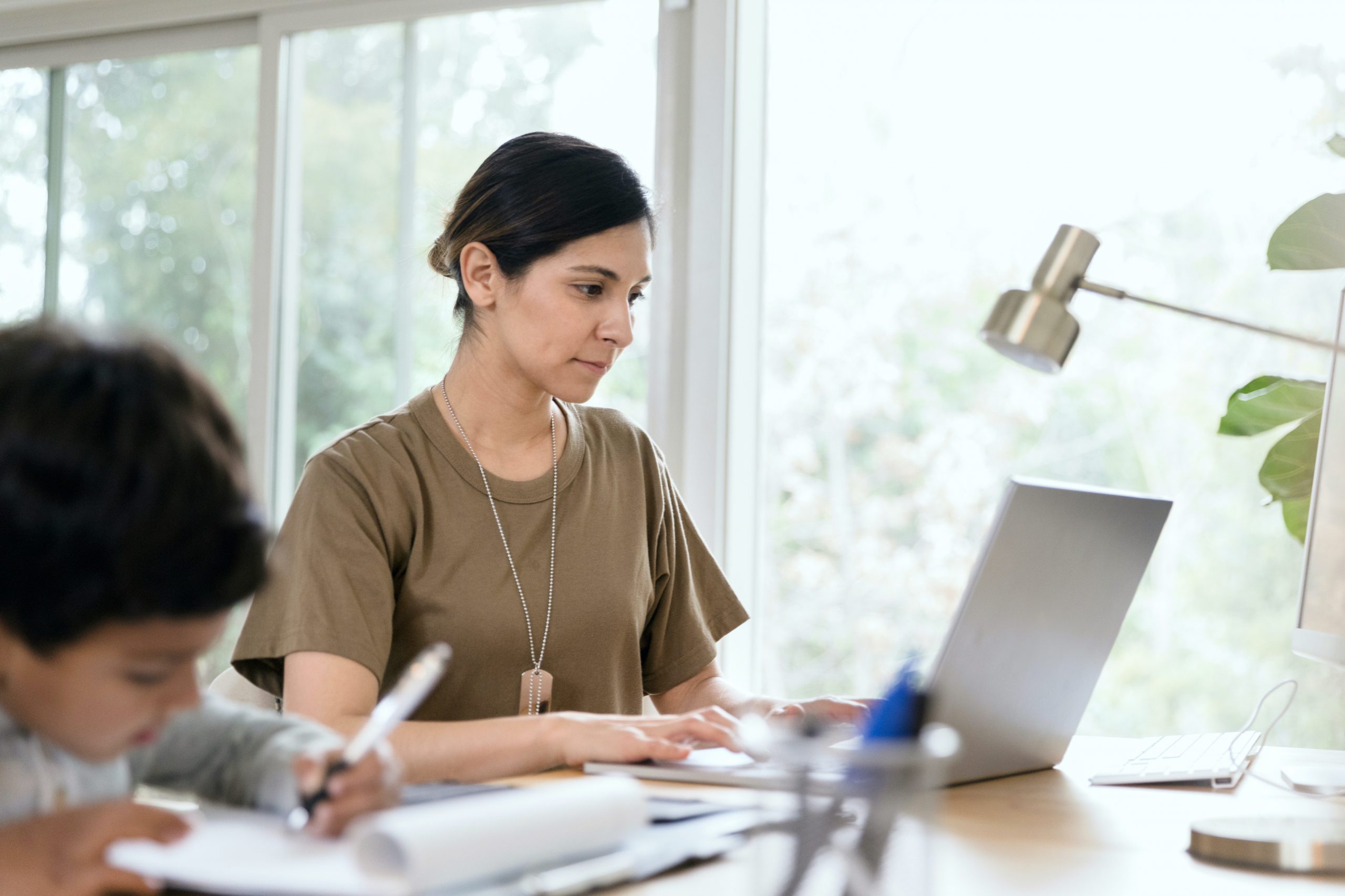 A mid adult female soldier concentrates while using a laptop in her home office. Her son is drawing a picture in the foreground.