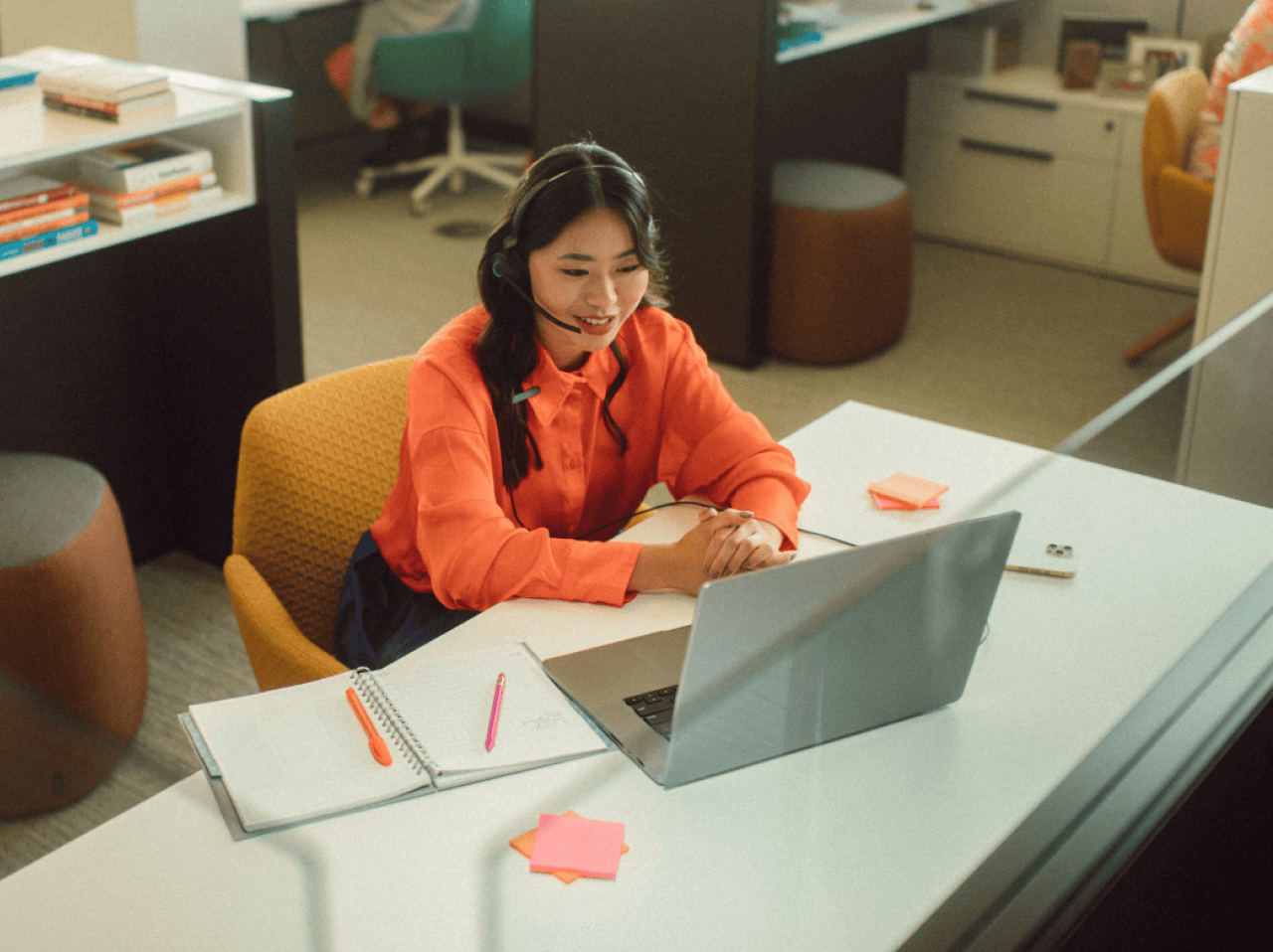 A woman working at an office desk, on a laptop, with a headset.