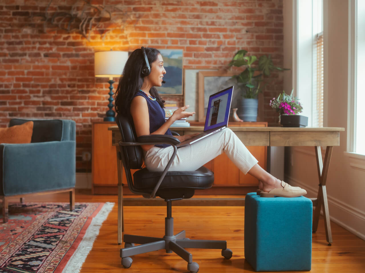 A woman working from home, in an office chair with a laptop on her lap and wearing a headset.