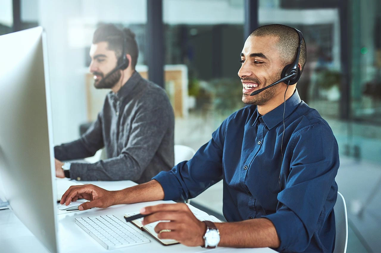 Photo of two individuals side by side with headsets on, at their workstations