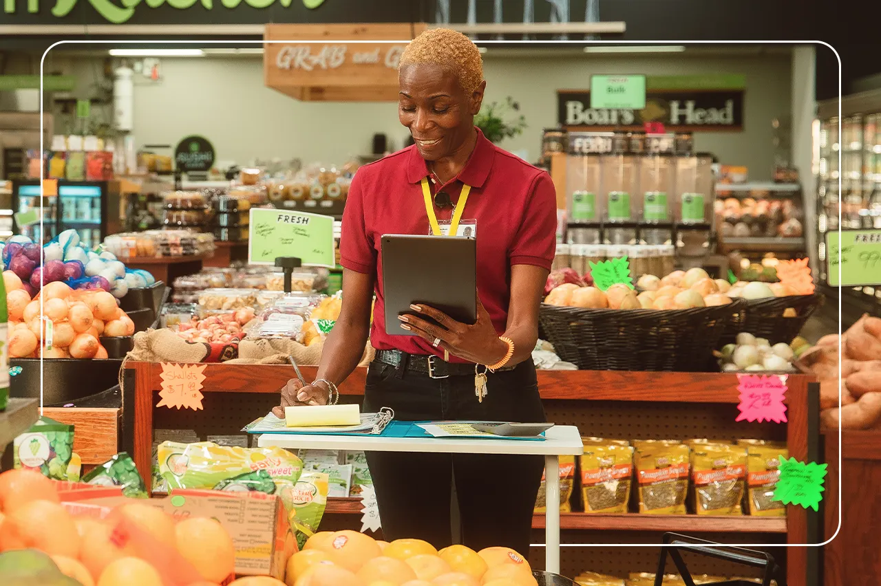 A smiling woman standing in a grocery store holding a tablet and writing in a notepad