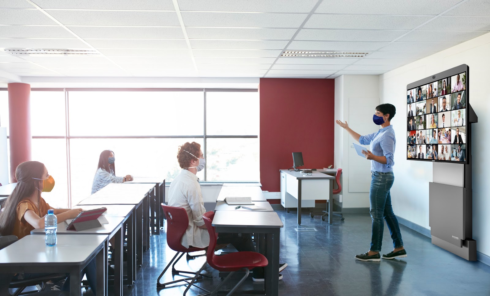 Teacher in front of class with Zoom gallery of students on screen behind her and students sitting at desks with masks on