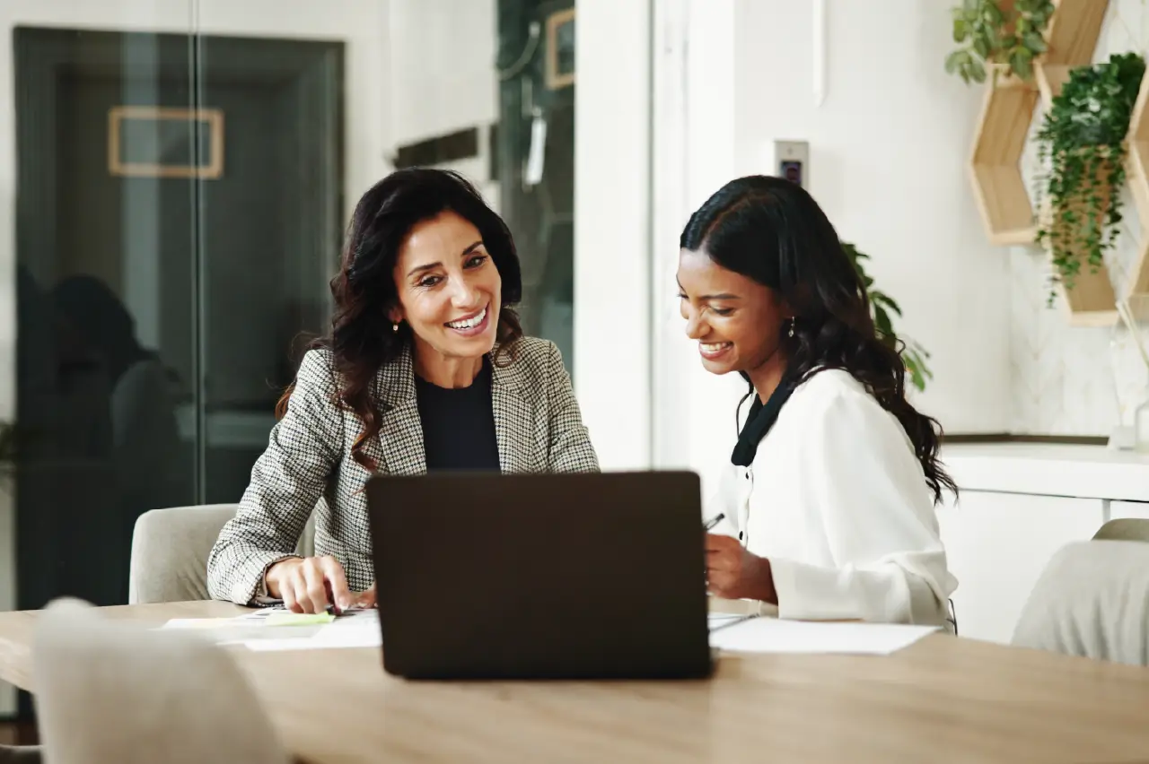 Two businesswomen sitting at a laptop, discussing task management strategies.