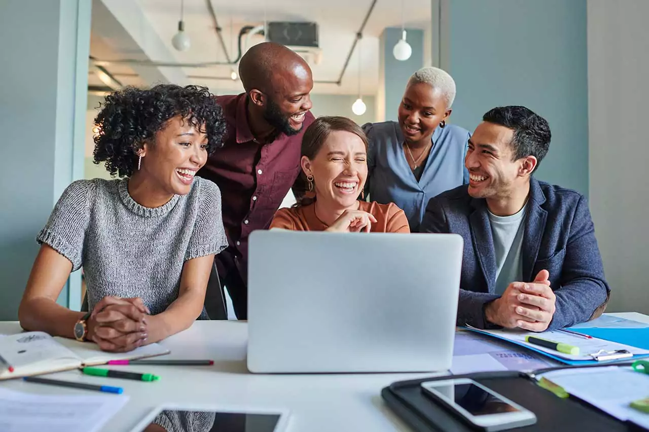 A team smiles and laughs around a laptop in an office setting